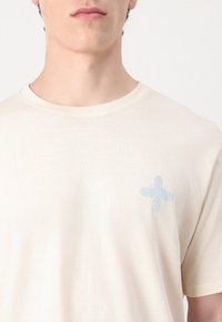 Camiseta color crema con cuello redondo y mangas cortas, que presenta un logo estampado en azul claro en la parte izquierda del pecho.