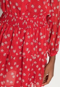 Vestido vermelho com bolinhas brancas e padrão floral. Apresenta cintura franzida e saia em camadas. Mangas longas com punhos elásticos. Tecido leve.