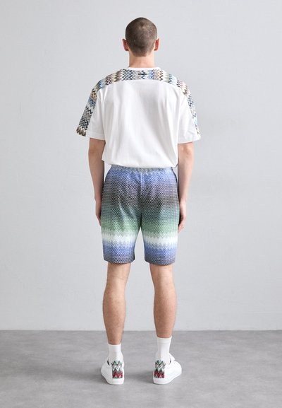 Herrenoutfit mit einem weißen T-Shirt mit Zickzackmuster an den Ärmeln, Shorts im blau-grünen Farbverlauf und weißen Sneakers mit passenden Akzenten.