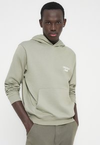 Νεαρός άνδρας με σκούρο δέρμα που φοράει ανοιχτό πράσινο φούτερ με κουκούλα και παντελόνι, τα χέρια στις τσέπες, στέκεται μπροστά σε ένα απλό λευκό φόντο.