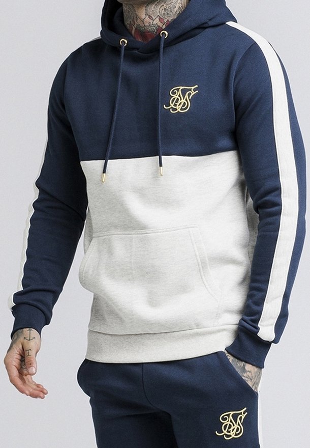Homme portant un sweat à capuche bicolore bleu marine et gris clair avec des bandes blanches sur les manches et un pantalon bleu marine assorti, tous deux avec un logo brodé doré.