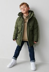 Giacca a vento in verde oliva scuro, con cappuccio, zip frontale e due tasche anteriori. Indossata sopra un hoodie beige e abbinata a jeans scuri e sneakers bianche.
