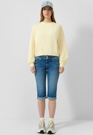 Jonge vrouw die een lichtgele sweatshirt draagt, opgerolde denim capri-jeans, witte sokken, sneakers en een lichte denim pet, staand tegen een grijze achtergrond.