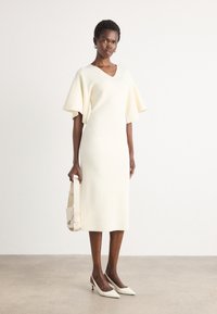 Cream knit dress with v-neck, short wide sleeves, and straight fit, paired with pointed white heels and a structured light bag. Smooth texture.