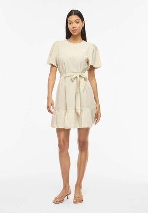 Femme portant une robe beige à manches courtes avec une ceinture nouée à la taille et un ourlet à volants, associée à des sandales à talons beiges, debout sur un fond blanc.