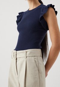 Kvinna som bär en marinblå ribbad ärmlös topp med volangärmar och högmidjade beige veckade byxor med bälte.