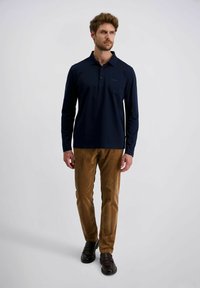 Man loopt, gekleed in een marineblauwe lange mouw polo, bruine corduroy broek en donker leren instappers tegen een effen lichte achtergrond.