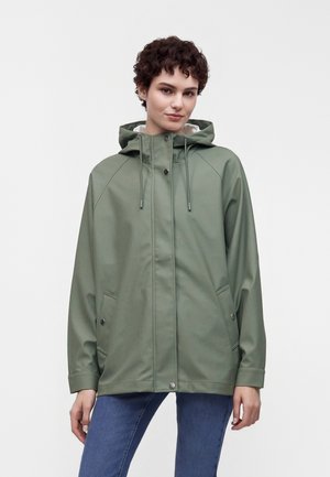 Jeune personne aux cheveux courts et bouclés portant une veste de pluie à capuche verte et un jean bleu, debout devant un fond uni.