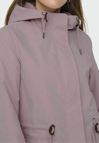 Kobieta w jasnofioletowej kurtce z kapturem, z regulowanymi sznurkami i zapięciem na suwak zakrytym klapą z przodu.