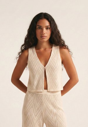 Mujer con cabello largo y rizado que lleva un chaleco de punto beige sin mangas y abierto al frente, junto con pantalones de talle alto a juego, sobre un fondo liso.