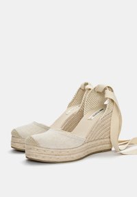 Beige espadrille wedges med vævede jute-såler, flade kanvas-overdele og bundet ankelremme. Textureret stof og rund tå-design.