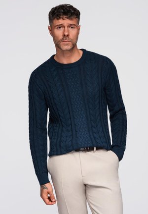 Pull en tricot bleu marine avec un motif en torsades, col rond et poignets côtelés. Associé à un pantalon de couleur claire.