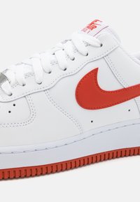 Bílá kožená teniska s červeným logem Nike, texturovanými bílými tkaničkami, polstrovaným límcem a červenou gumovou podrážkou s drážkovaným vzorkem pro lepší trakci.