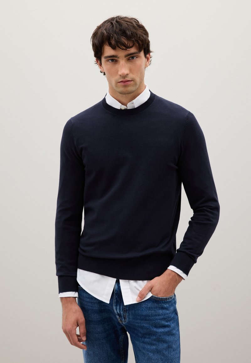 Maglione lavorato a maglia blu navy con maniche lunghe, scollo rotondo e polsini a coste, indossato sopra una camicia bianca con colletto. Jeans in denim casual.
