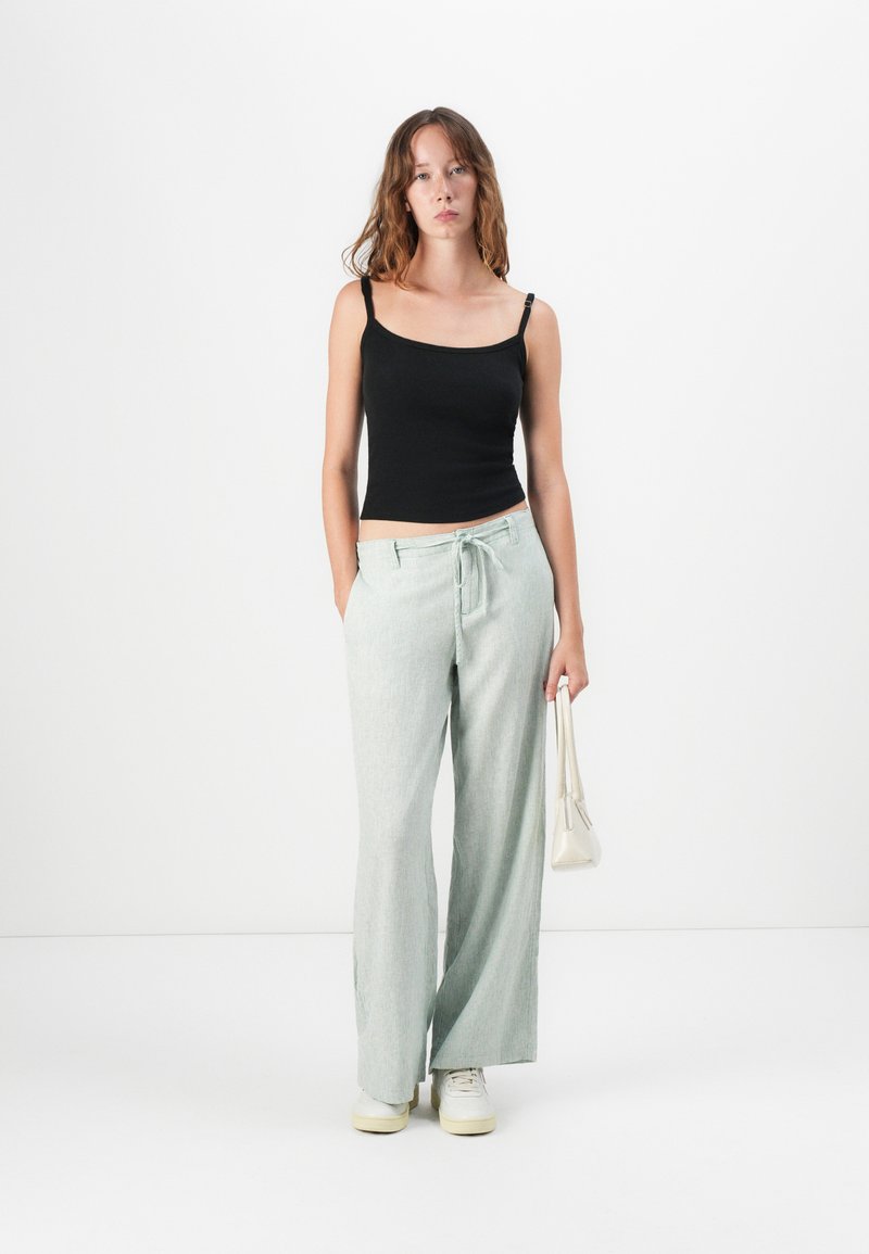 Czarna dopasowana koszulka na ramiączkach, zestawiona z szerokimi, jasnoblokowymi spodniami o fakturowanym wzorze i wiązaniu w talii. Trzyma biały torebkę.