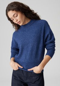 Maglione in lana blu navy con collo alto, polsini e orlo a coste. Tessuto morbido, vestibilità comoda, caratterizzato da maniche larghe e cadenti. Abbinato a jeans scuri.