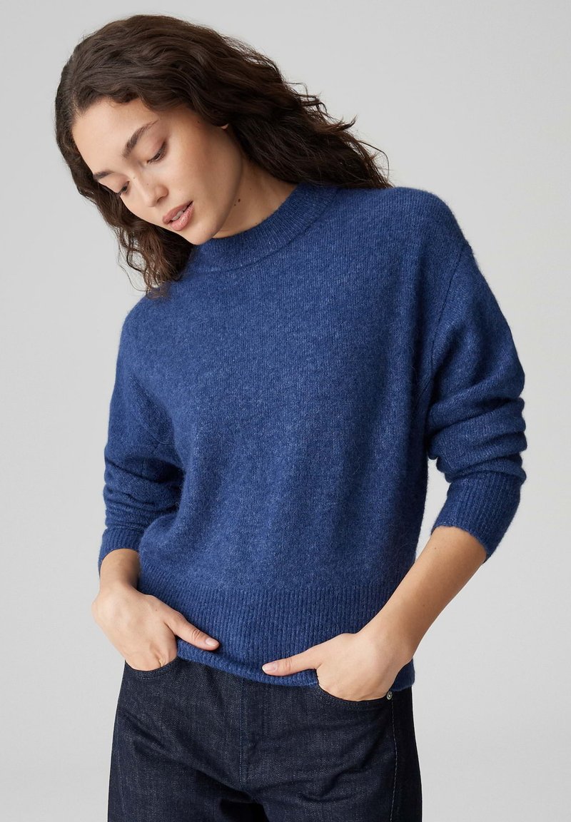 Maglione in lana blu navy con collo alto, polsini e orlo a coste. Tessuto morbido, vestibilità comoda, caratterizzato da maniche larghe e cadenti. Abbinato a jeans scuri.