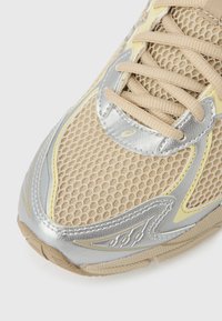 Ténis atlético bege com parte superior em malha, detalhes em prata e amarelo. Possui atacadores e sola texturizada para aderência e suporte.