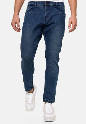 Jeans in denim blu scuro con vestibilità slim, dotati di cinque tasche, chiusura con bottone e dettagli di cucitura sottili. Indossati con sneakers bianche.