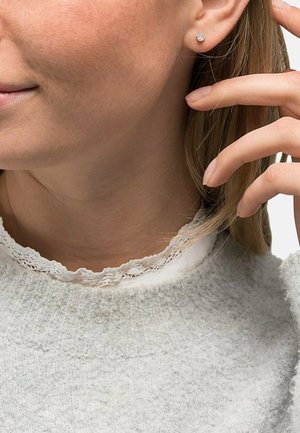 Silberne Ohrringe mit Blütenmuster, die von einem Model getragen werden. Ein weicher grauer Pullover mit einer Spitzenkragen und strukturiertem Stoff ist sichtbar.
