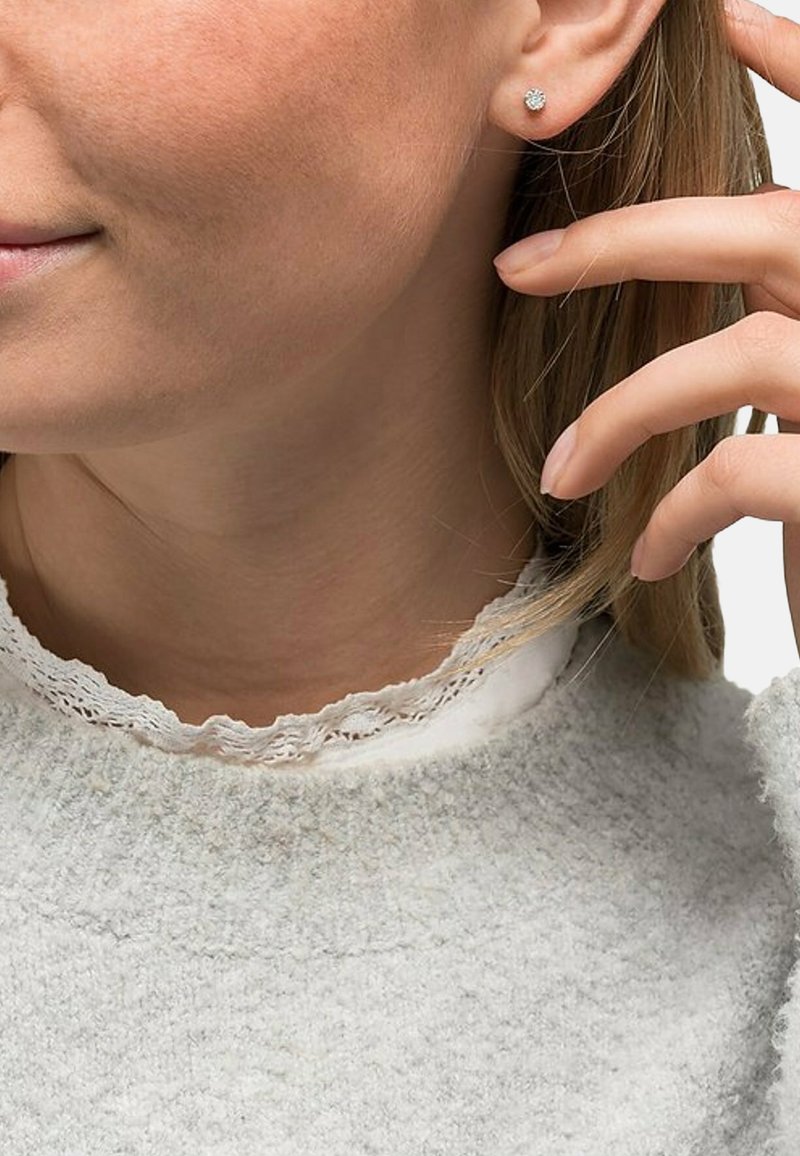 Silberne Ohrringe mit Blütenmuster, die von einem Model getragen werden. Ein weicher grauer Pullover mit einer Spitzenkragen und strukturiertem Stoff ist sichtbar.
