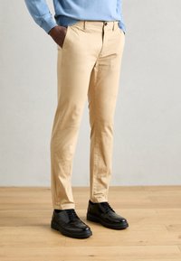 Beige katoenen broeken met een slim fit, voorzien van zijzakken en een knoopsluiting, gecombineerd met zwarte leren schoenen met veters en een dikke zool.