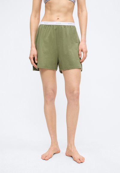 Vue de face d'une personne portant un short à taille élastique vert olive avec une ceinture blanche de marque, debout pieds nus sur un fond clair uni.