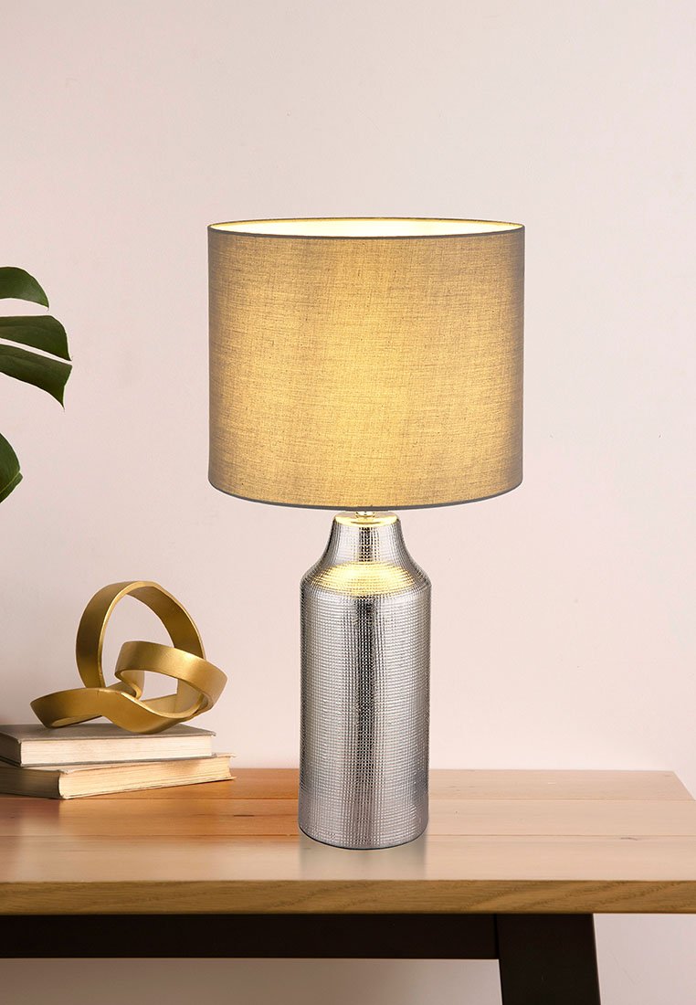 Stolní lampa s texturovanou stříbrnou základnou a kulatým, světlým béžovým látkovým stínidlem. Umístěná na dřevěném stole vedle dekorativních předmětů.