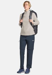 Donna che indossa un fleece beige con zip, pantaloni da escursionismo blu scuro, sneakers navy e uno zaino nero, in piedi davanti a uno sfondo chiaro e semplice.