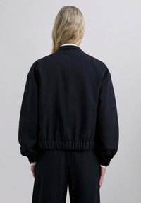 Persona con cabello largo rubio viste una chaqueta negra con rayas y pantalones a juego, de pie frente a un fondo gris claro liso.