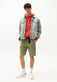 Chaqueta de denim sobre un hoodie naranja, pantalones cortos de cargo oliva, zapatillas blancas con detalles en negro y calcetines cortos blancos, de pie contra un fondo liso.