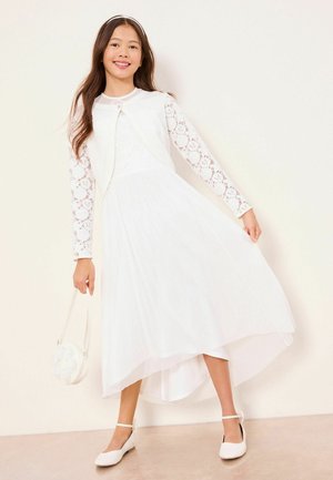 Mädchen in einem weißen Kleid mit Spitzenärmeln und einer Strickjacke hält eine runde Handtasche, trägt weiße Schuhe mit Knöchelriemchen und lächelt vor einem schlichten Hintergrund.