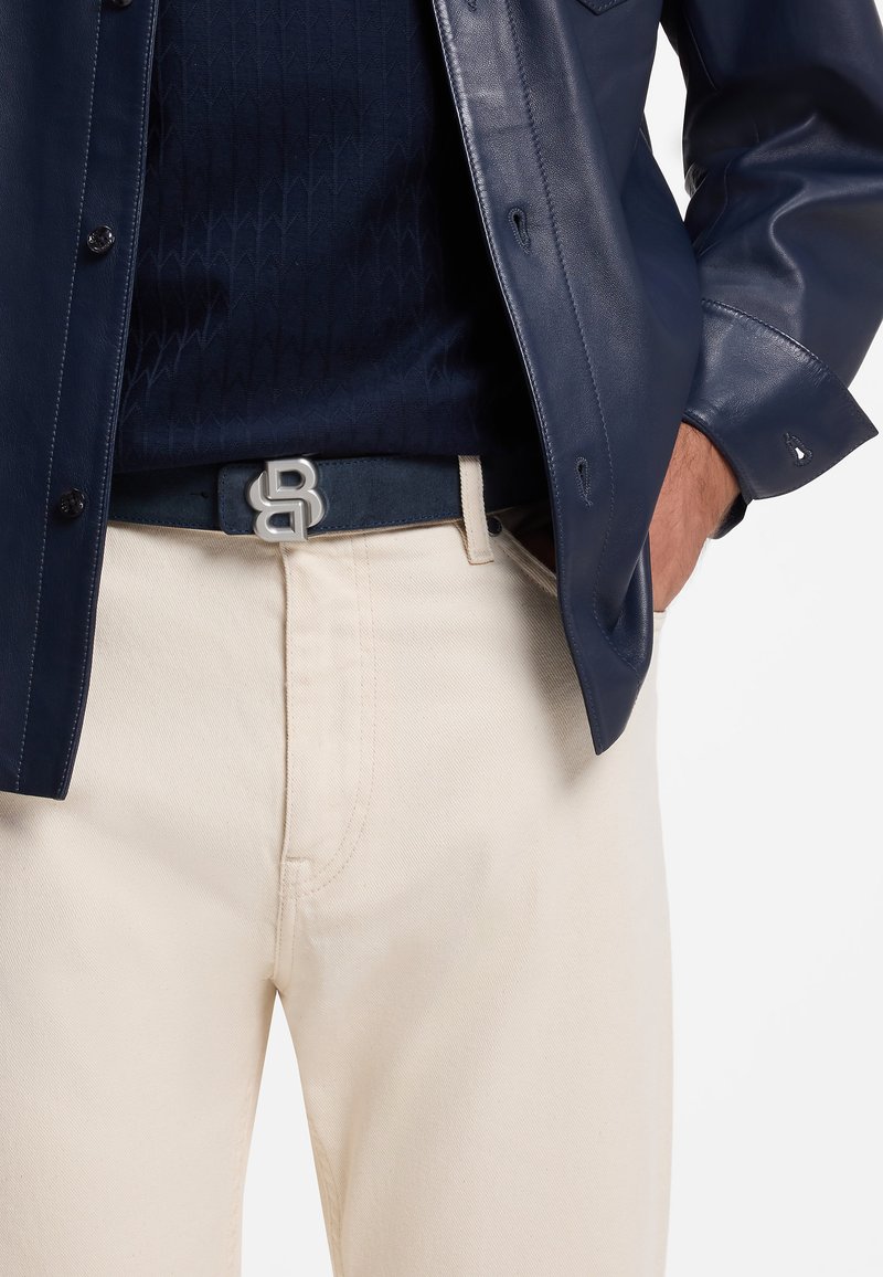 Homme portant un pantalon crème, une ceinture bleu marine avec boucle argentée, une chemise texturée bleu marine et une veste en cuir bleu marine avec la main dans la poche.