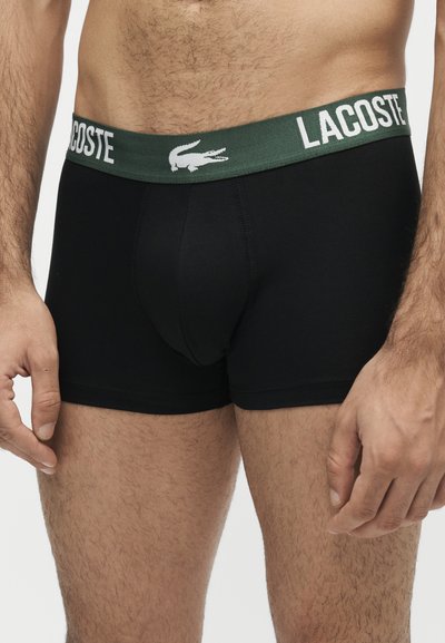 Мъж, носещ черни боксерки с зелен колан с логото на Lacoste и бял текст на едноцветен фон.