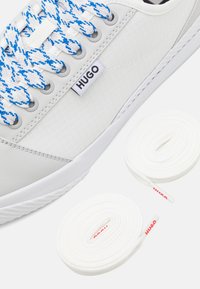 Sneakers blanches avec des lacets rayés bleus et une étiquette de logo "HUGO", accompagnées de deux lacets de remplacement blancs enroulés.