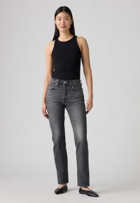 Zwarte mouwloze bodysuit gecombineerd met hooggesneden grijze jeans met een rechte pijp, subtiele vervaging en zwarte flats. Neutrale achtergrond.