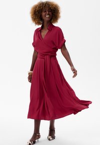 Rode wikkeljurk met korte mouwen, strik taille, vloeiende rok en lichtgewicht stof. Gestyled met patronen hakken en grote ronde oorringen.