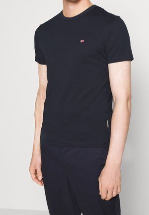 Homme portant un t-shirt à manches courtes bleu marine uni avec un petit emblème de drapeau rouge et blanc sur la poitrine, ainsi qu'un pantalon assorti de couleur foncée.