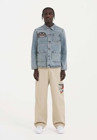 Vaaleansininen farkkutakki, jossa on kaksi etutaskua ja brodeerattu design. Beige leveälahkeiset housut, joissa on värikäs graafinen yksityiskohta, yhdistettynä valkoisiin tennareihin.