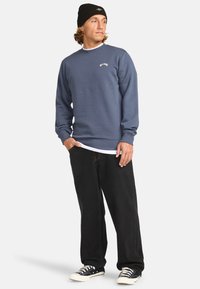 Sudadera azul con puños y dobladillo de canalé, que presenta un pequeño logo blanco en el pecho. Combinada con pantalones anchos negros y zapatillas.