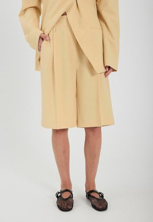 Svetlo rumeni krojeni kratki hlače s ravnim krojem, gubami spredaj in dvema stranskima žepoma. V kombinaciji z črnimi športnimi čevlji z mrežastimi trakovi.