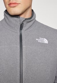 Grå fleece zip-up jakke med høj krave, sort lynlås og broderet logo på den øverste venstre brystområde. Blød tekstur.