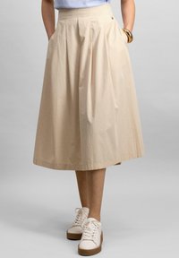Beige cotton midi skirt with pleats, curved hem, and side pockets, paired with white sneakers featuring a thick brown sole.