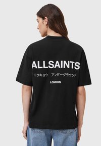 Čierne voľné tričko z bavlny s výrazným bielym nápisom na chrbte: "ALLSAINTS," jazyky a "LONDON." Krátke rukávy, uvoľnený strih.
