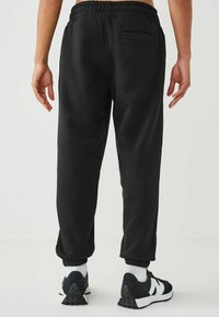 Pantalones de chándal negros con cintura elástica y perneras ajustadas, que cuentan con un bolsillo trasero y una tela suave y texturizada. Llevados con zapatillas negras.