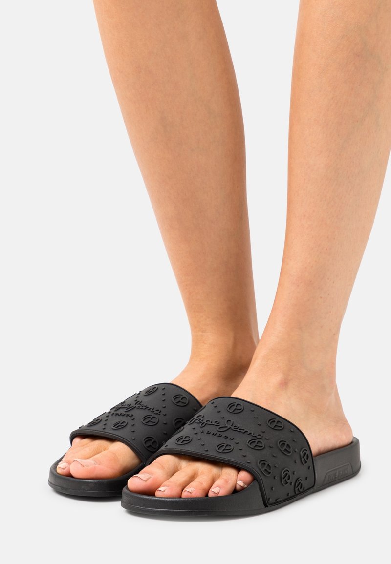 Pieds portant des sandales noires à lanières ornées de logos en relief et du nom de la marque sur les brides supérieures texturées, sur un fond uni.