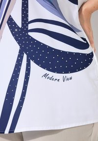 Blusa blanca con un diseño abstracto en azul marino, adornada con piedras de strass y el texto "Vista Moderna" cerca del dobladillo. Textura suave, corte holgado.