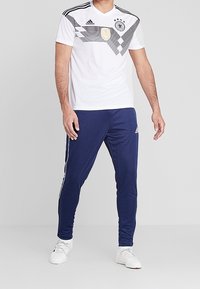 Man som bär en vit Tyskland fotbollströja och navy Adidas träningsoverallbyxor med vita sneakers som står mot en grå bakgrund.