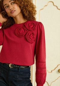 Červená mikina s trojicou textúrovaných, látkových aplikácií v tvare ruží na prednej strane, s dlhými rukávmi a dekoratívnymi výrezmi v blízkosti manžiet.