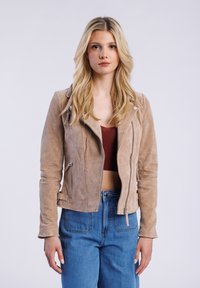 Tan suede motorjack met een rits aan de voorkant, zijzakken en een reverskraag. Gecombineerd met hooggenaaide blauwe spijkerbroek. Gladde textuur.